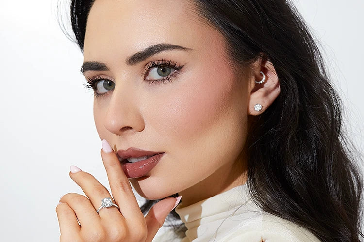 Woman with long dark hair and flawless makeup wearing a sleeveless cream top, diamond stud earrings, and a large diamond ring, posing with her hand gently touching her lips.