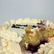 Close-up of a partially eaten white frosted cake with piped decorations and dark berry filling visible inside, revealing a yellow sponge interior.
