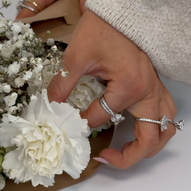 Close-up of a hand arranging white carnations and baby's breath flowers, adorned with multiple diamond rings in various styles including solitaire and pavé settings.
