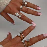 Close-up of hands with light pink manicured nails wearing multiple diamond rings in various shapes and settings, including solitaire, halo, and pavé bands on both white and yellow gold bands.