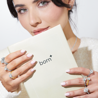 Woman with pink manicured nails holding a "born" booklet, showcasing multiple diamond rings in white gold, yellow gold, and platinum on both hands. She wears soft makeup and a white textured top.