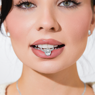 Close-up of a woman with flawless makeup and diamond stud earrings, holding an oval-cut diamond halo engagement ring between her teeth, smiling slightly.