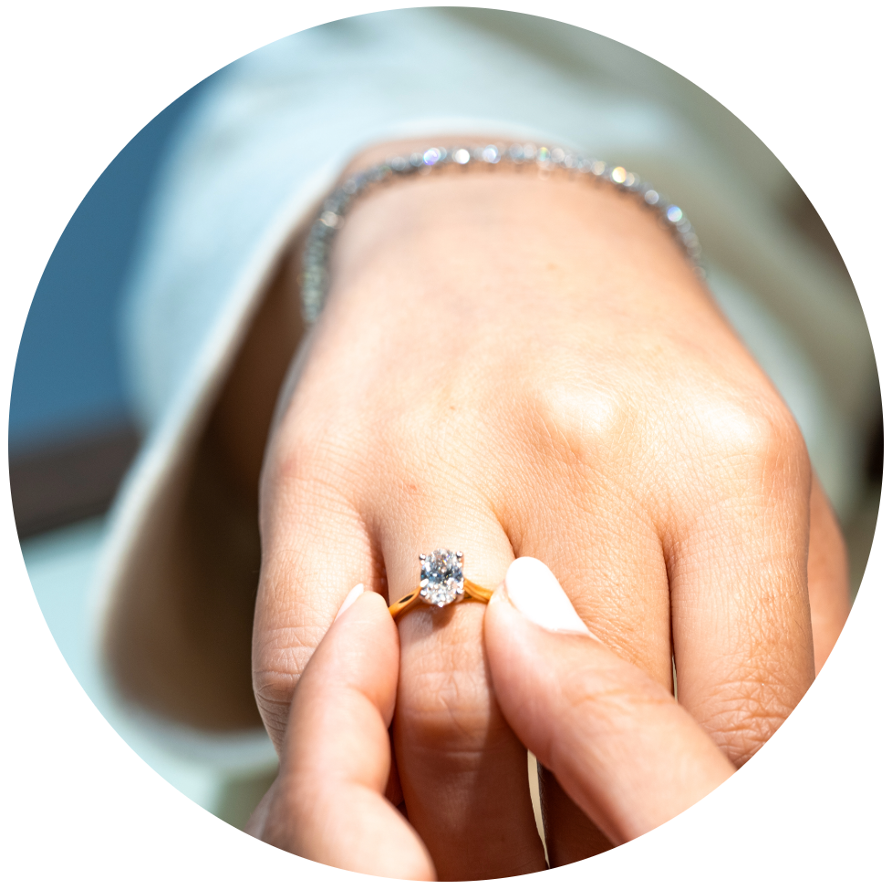 Close-up of hands gently placing an oval-cut diamond solitaire engagement ring set on a polished yellow gold band onto a finger, complemented by a diamond bracelet.