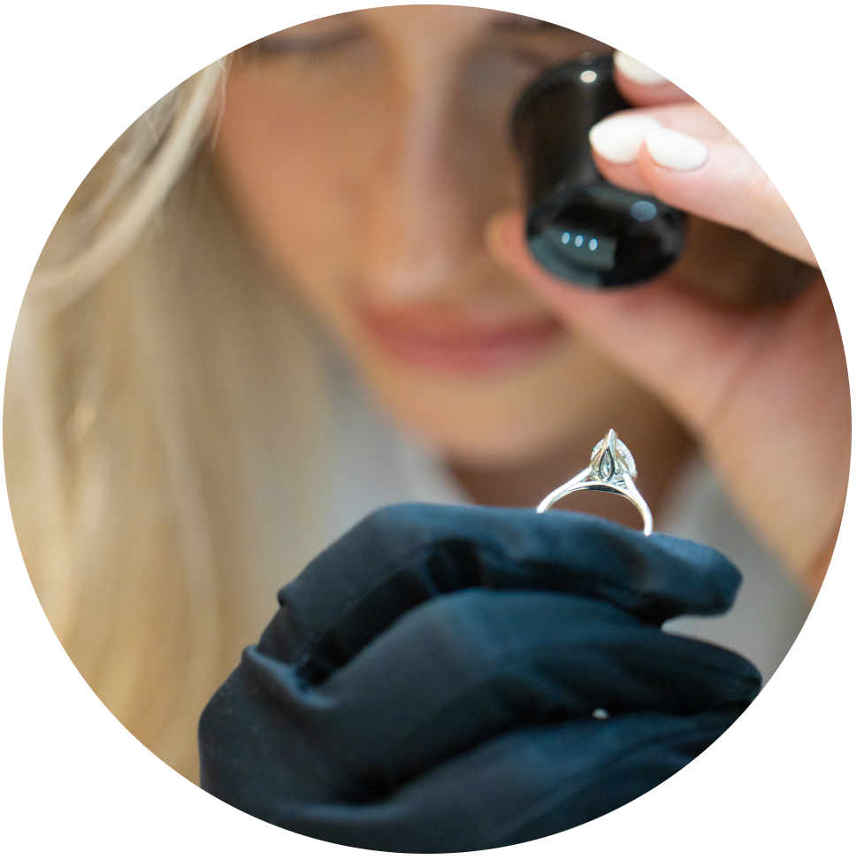 Jeweller closely examining a diamond engagement ring through a loupe, wearing black gloves, focusing carefully on the ring's detailed setting.