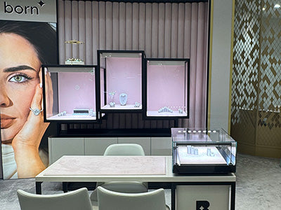 Jewellery display area featuring a "born" branding poster with a woman wearing diamond jewellery, three pink-lit glass display cases with various pieces, a modern counter with seating, and an additional display box on the counter.